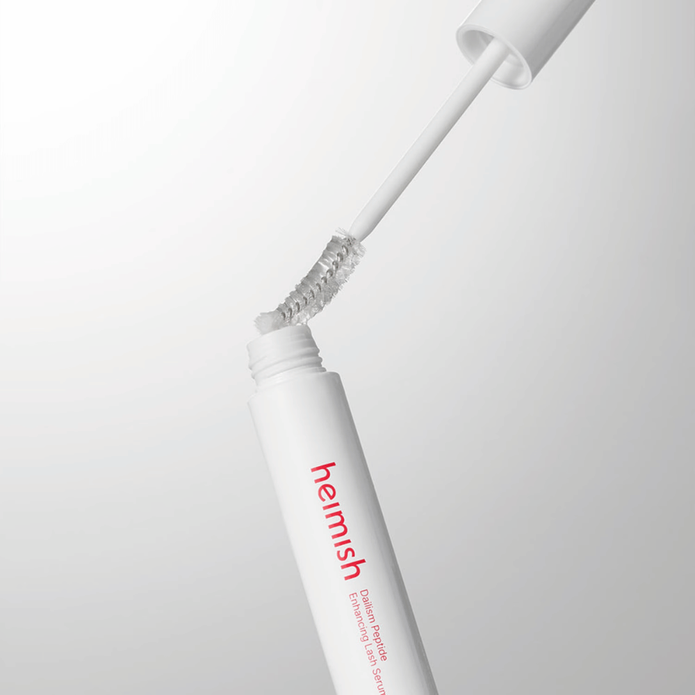 Κοντινό πλάνο του απλικατέρ του Heimish Dailism Peptide Enhancing Lash Serum. Το βουρτσάκι τραβιέται έξω από το λευκό δοχείο και παρουσιάζει τη διαφανή σύνθεση του ορού που εφαρμόζεται στις βλεφαρίδες.