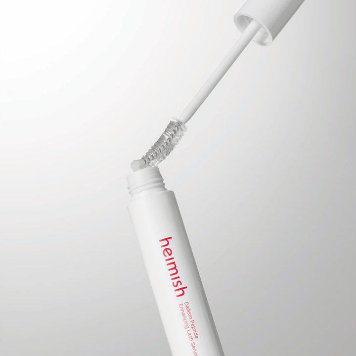 Κοντινό πλάνο του απλικατέρ του Heimish Dailism Peptide Enhancing Lash Serum. Το βουρτσάκι τραβιέται έξω από το λευκό δοχείο και παρουσιάζει τη διαφανή σύνθεση του ορού που εφαρμόζεται στις βλεφαρίδες.