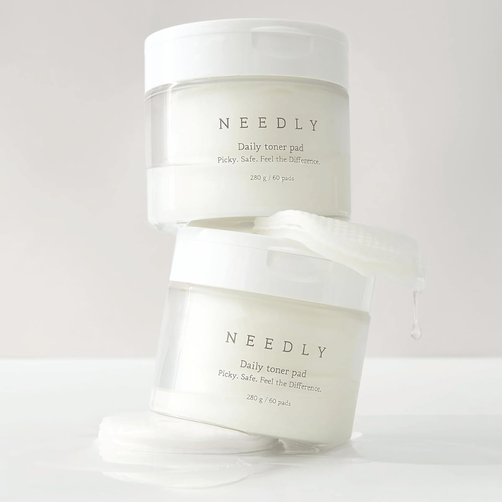 Δύο βάζα με Needly Daily Toner Pad στοιβαγμένα το ένα πάνω στο άλλο. Τα βάζα είναι λευκά με μινιμαλιστικό κείμενο και περιέχουν 60 τονέρ παντς το καθένα. Ένα τονέρ παντ βρίσκεται μερικώς έξω από το πάνω βάζο με υγρό που στάζει.