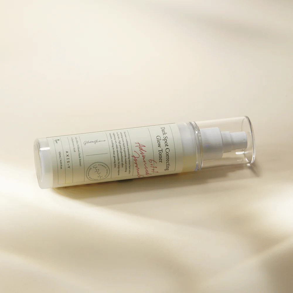 Ένα ξαπλωμένο μπουκάλι του "AXIS-Y Dark Spot Correcting Glow Toner" τοποθετημένο σε μια φωτεινή επιφάνεια. Το μπουκάλι είναι διάφανο με μια λευκή ετικέτα και έχει μια λευκή αντλία σπρέι. Η ετικέτα δείχνει πληροφορίες για τις φωτεινές και ενυδατικές ιδιότητες του προϊόντος και περιέχει βασικά συστατικά όπως Γλουταθειόνη και Νιασιναμίδη.