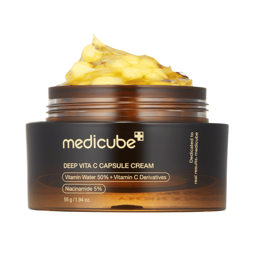 Μια κοντινή λήψη του "Medicube Deep Vita C Capsule Cream", που δείχνει την υφή της κρέμας και τις κάψουλες βιταμίνης C. Η συσκευασία είναι σκούρο καφέ με χρυσά γράμματα και περιγράφει το περιεχόμενο του νερού βιταμινών (50%) και της νιασιναμίδης (5%), παρέχοντας μια οπτική παρουσίαση των ενυδατικών και βελτιωτικών ιδιοτήτων της επιδερμίδας του προϊόντος.