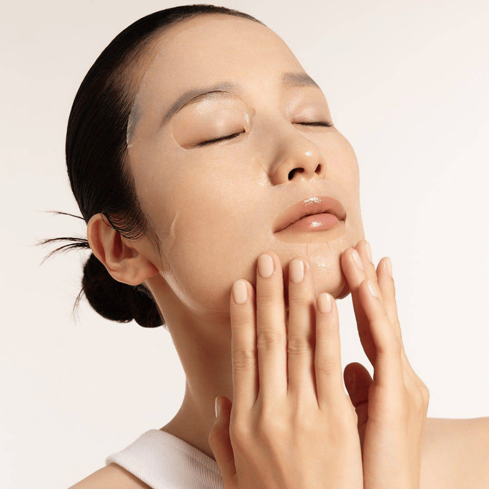 Μοντέλο με ενυδατωμένο και λαμπερό δέρμα εφαρμόζει μάσκα φύλλου, εστίαση στη λάμψη, ομοιόμορφο τόνο δέρματος και λάμψη.