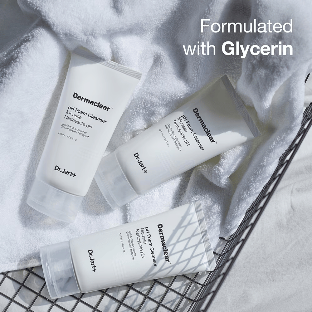 Τρεις σωλήνες του Dr.Jart+ Dermaclear pH Foam Cleanser βρίσκονται πάνω σε μια λευκή πετσέτα φροτέ σε ένα μεταλλικό καλάθι, με το κείμενο "Formulated with Glycerin" στην πάνω γωνία.