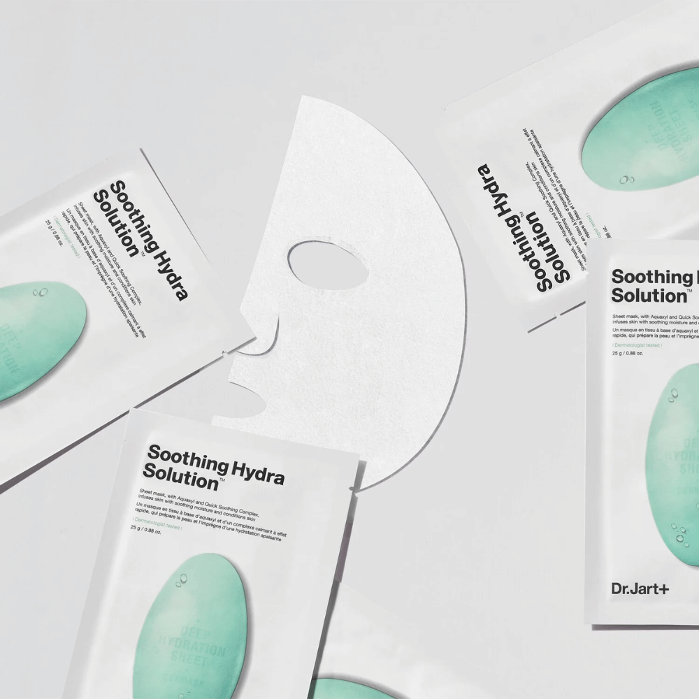 Πολλές συσκευασίες από μάσκες προσώπου "Soothing Hydra Solution" από την Dr.Jart+ είναι απλωμένες σε μια φωτεινή επιφάνεια. Μια ανοιγμένη μάσκα τοποθετείται στη μέση, δείχνοντας τη λευκή και διάτρητη δομή της.