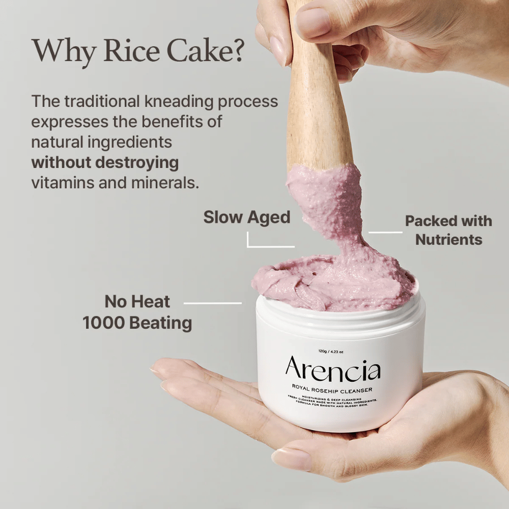 Ένα χέρι κρατά ένα ανοιχτό βάζο του Arencia Royal Rosehip Cleanser, με μια ροζ, καθαριστική φόρμουλα με υφή μοτσί που σηκώνεται με ξύλινη σπάτουλα – δείχνει την υφή του προϊόντος και την παραδοσιακή μέθοδο παρασκευής.