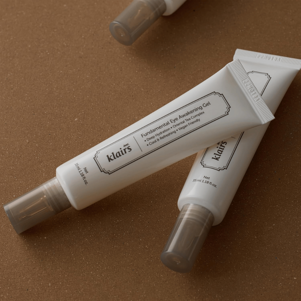 Lifestyle εικόνα με δύο σωληνάρια του Klairs Fundamental Eye Awakening Gel τοποθετημένα σε ζεστό καφέ φόντο. Η σύνθεση αναδεικνύει τον κομψό σχεδιασμό του προϊόντος και την αίσθηση πολυτέλειας, ενώ παράλληλα εστιάζει στη φροντίδα των ματιών και την ενυδάτωση.