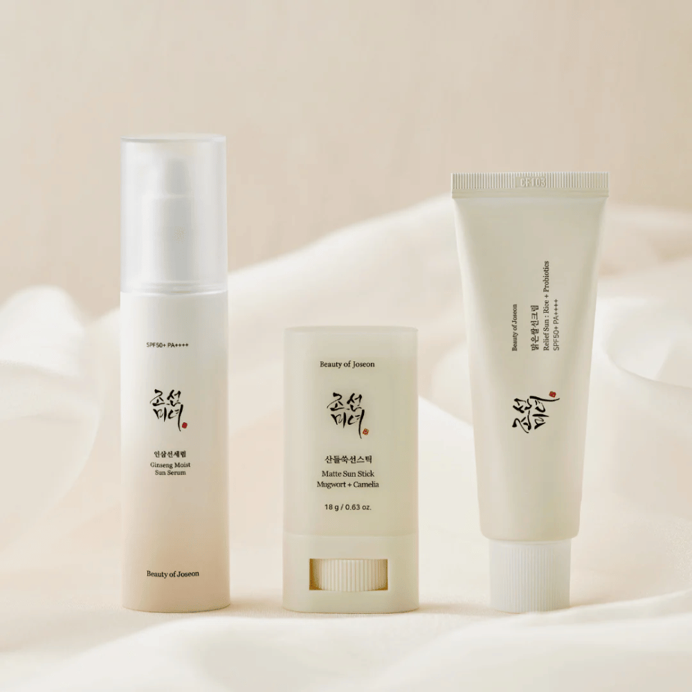 Τρία αντηλιακά προϊόντα από Beauty of Joseon - Ginseng Moist Sun Serum, Matte Sun Stick και Relief Sun Rice + Probiotics - είναι παραταγμένα σε ένα κρεμ ύφασμα με απαλό φως.