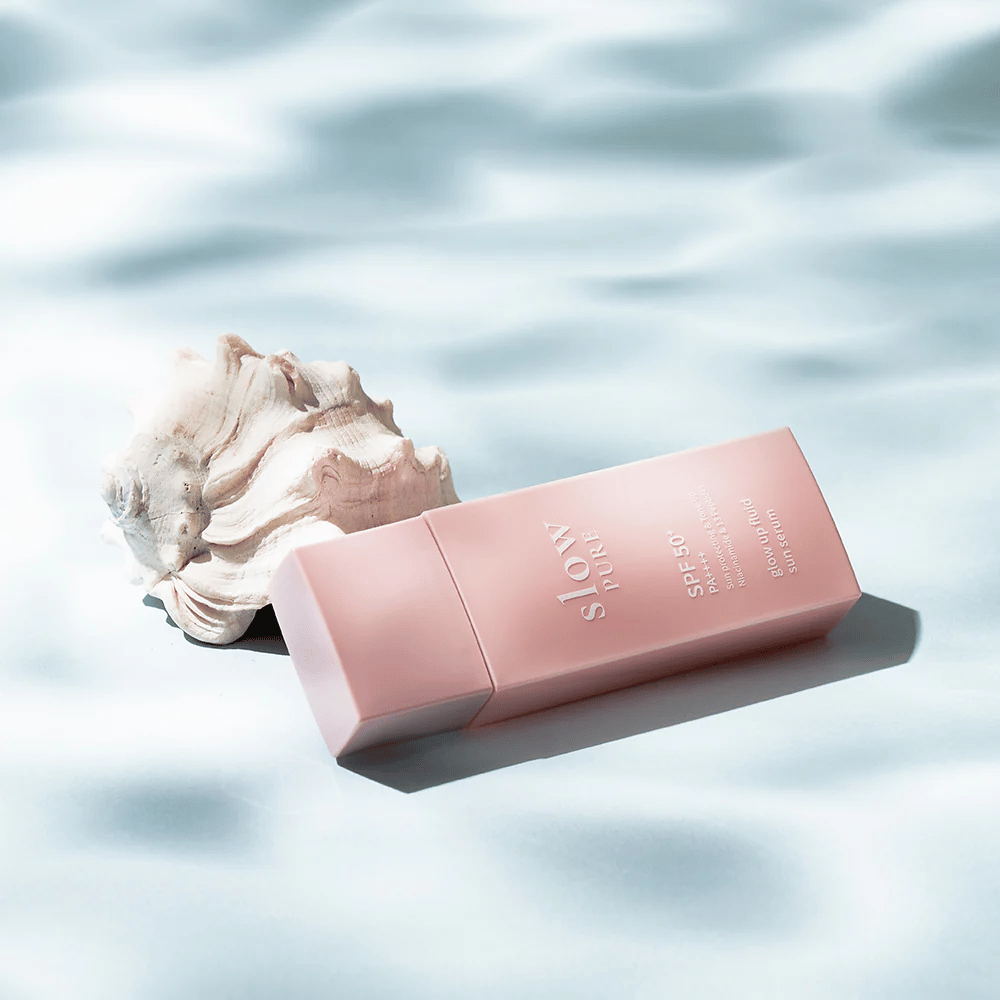 Slow Pure Glow Up Fluid Sun Serum δίπλα σε κοχύλι και εφέ νερού, τονίζει την αντηλιακή προστασία και τη λάμψη.