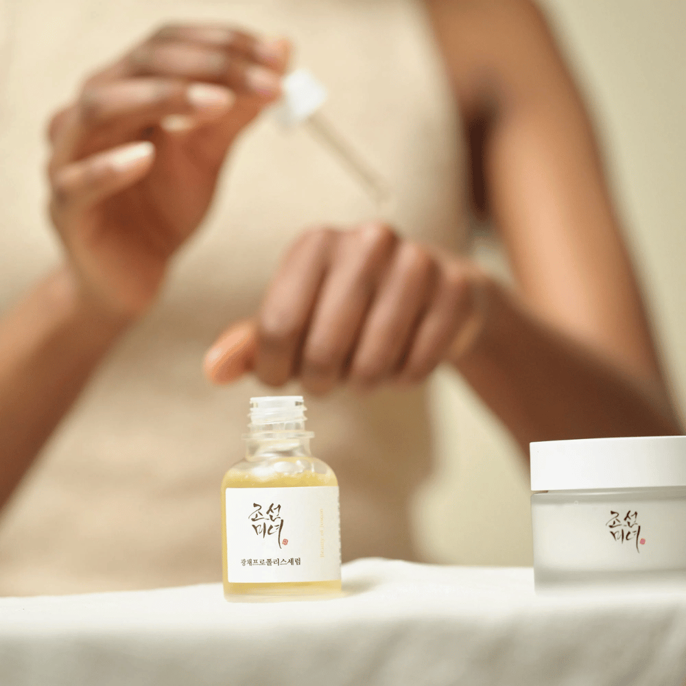 Άτομο που εφαρμόζει Beauty of Joseon Glow Serum με πιπέτα από γυάλινο μπουκάλι, με ένα βάζο της ίδιας σειράς δίπλα στο τραπέζι.