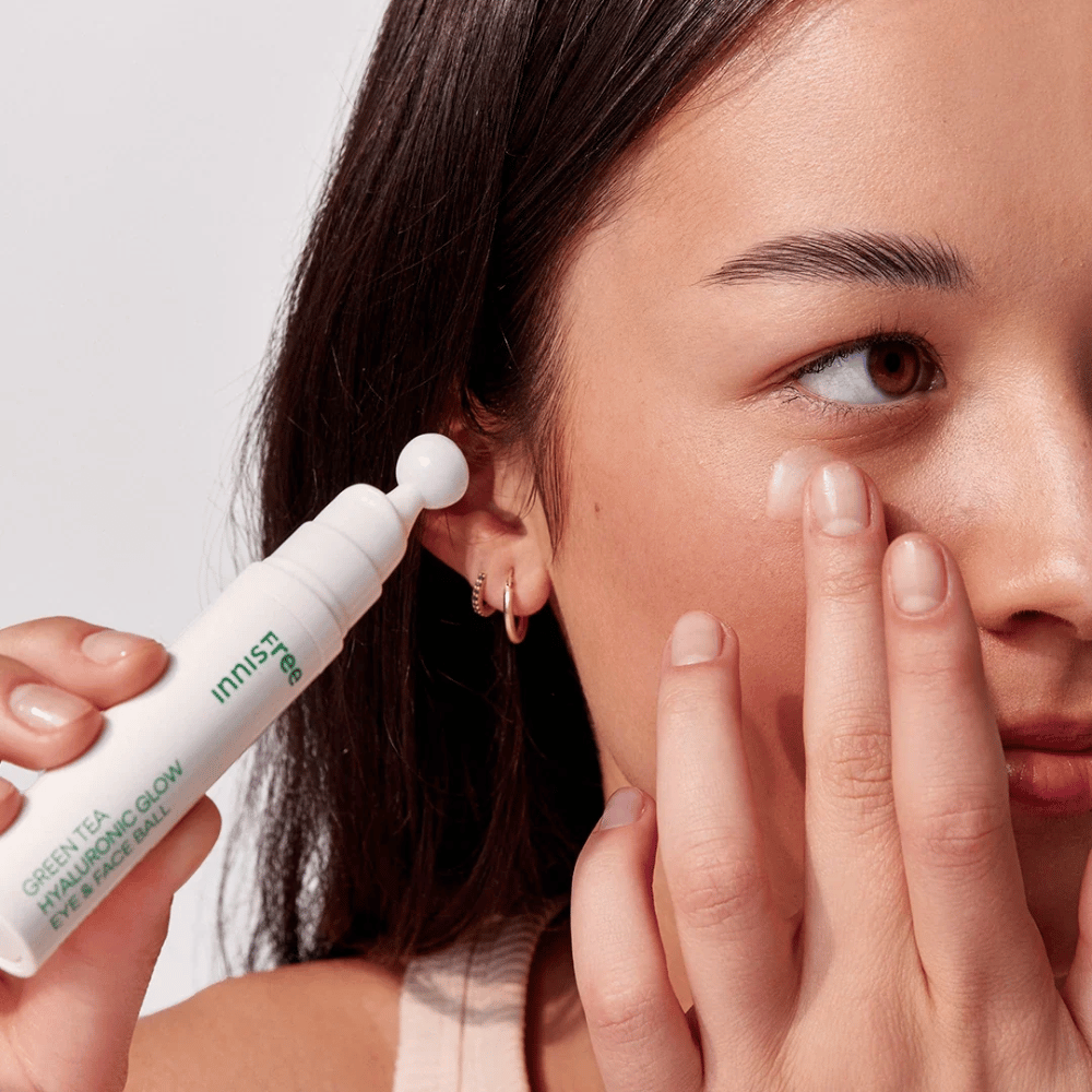 Ένα άτομο εφαρμόζει Innisfree Green Tea Hyaluronic Glow Eye & Face Ball στο δέρμα του κάτω από τα μάτια. Το προϊόν έχει ένα λευκό, στρογγυλό μέρος εφαρμογής και είναι σε ένα λευκό μπουκάλι με πράσινο κείμενο. Το πρόσωπο του χρήστη φαίνεται μερικώς και εστιάζει στην εφαρμογή του προϊόντος.