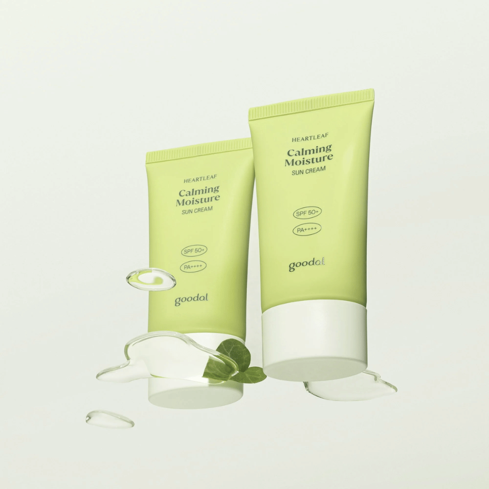 Δύο σωλήνες Goodal Heartleaf Calming Moisture Sun Cream SPF50+ PA++++ περιτριγυρισμένοι από φύλλα φυτών και σταγόνες ορού σε φωτεινό φόντο.