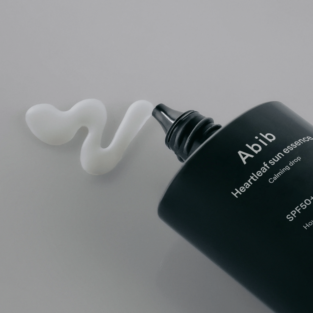Κοντινή εικόνα του Abib Heartleaf Sun Essence Calming Drop με την αντηλιακή κρέμα εκτεθειμένη σε μια λευκή, κυματιστή γραμμή σε μια φωτεινή επιφάνεια.