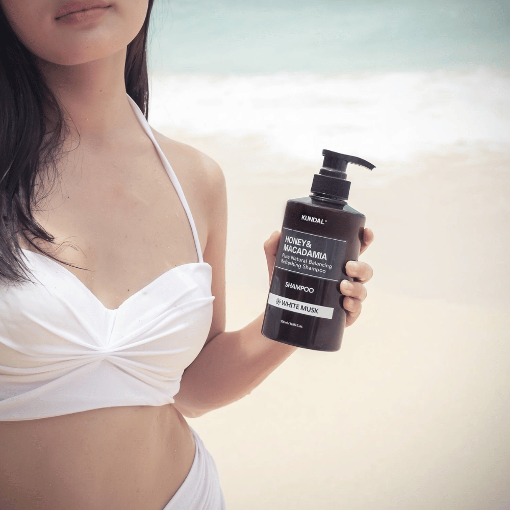Γυναίκα με λευκό μπικίνι κρατάει KUNDAL Honey & Macadamia Nature Shampoo White Musk σε μια παραλία με τη θάλασσα στο φόντο.