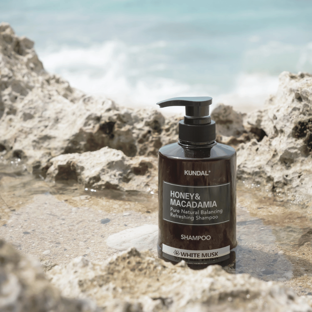 KUNDAL Honey & Macadamia Nature Shampoo White Musk τοποθετημένο σε βράχια δίπλα στη θάλασσα, ένα φυσικό προϊόν περιποίησης μαλλιών με φρέσκο άρωμα.
