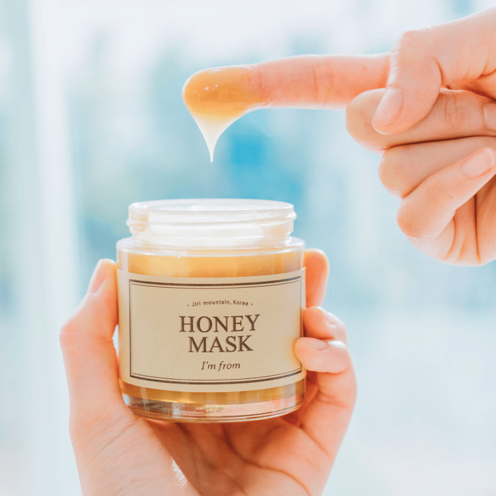 Ένα χέρι κρατάει ανοιχτό βάζο του "I'm from Honey Mask", και ένα δάχτυλο βυθίζεται στη χρυσή μάσκα σε ανοιχτό φόντο.