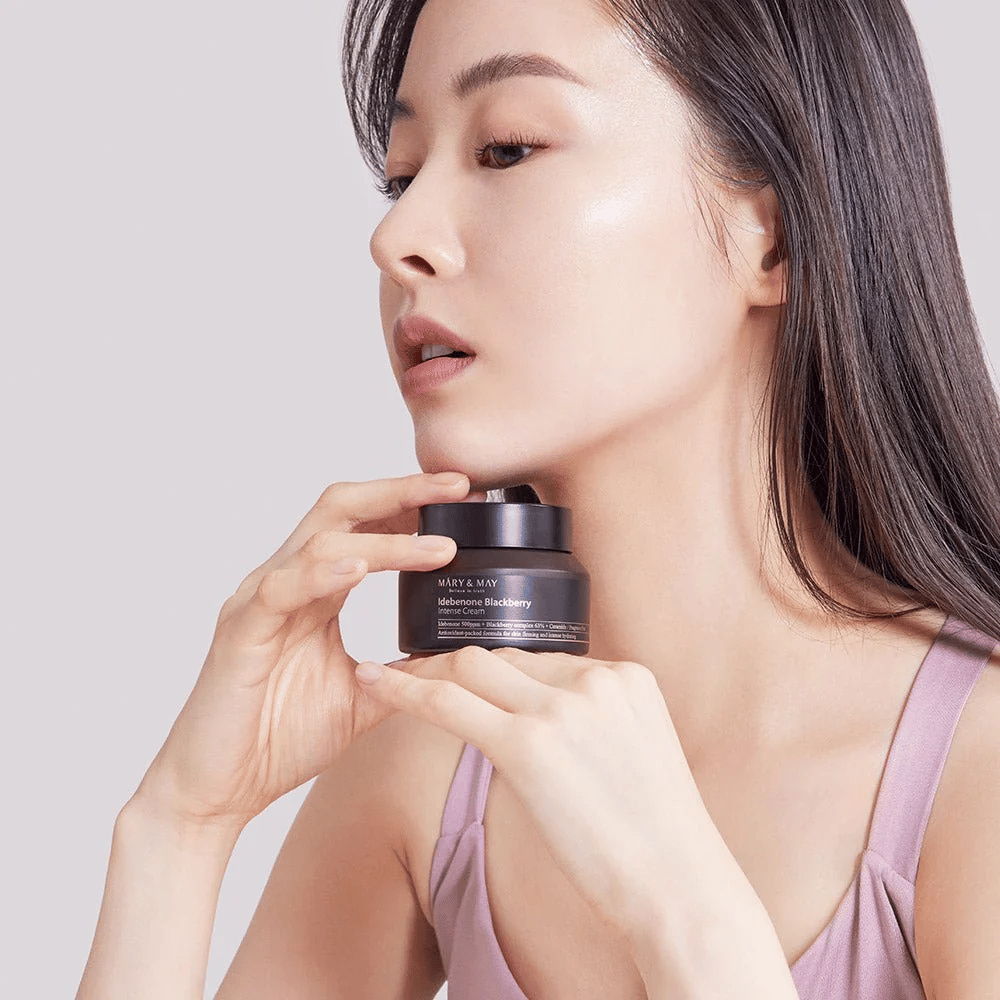 Μια γυναίκα κρατάει ένα δοχείο με MARY & MAY Idebenone Blackberry Intense Cream. Έχει λαμπερή επιδερμίδα και χαμογελά ελαφρά, ντυμένη με ένα αμάνικο τοπ. Το προϊόν είναι μια εντατική κρέμα με idebenon και σύνθετο βατόμουρου που προορίζεται για τη σφριγηλότητα και την ενυδάτωση του δέρματος.