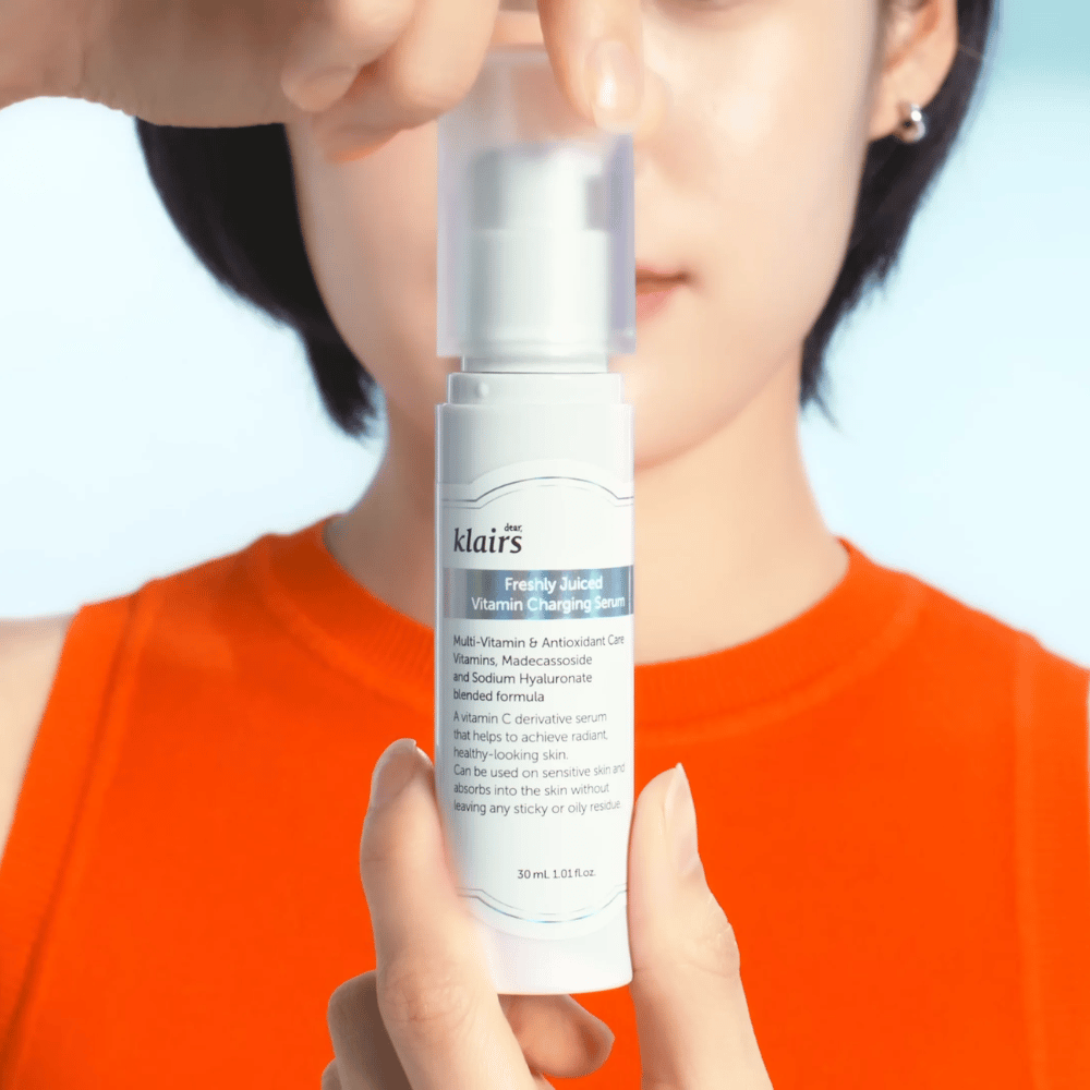 Γυναίκα με πορτοκαλί μπλούζα κρατάει Klairs Freshly Juiced Vitamin Charging Serum μπροστά της σε μπλε φόντο.