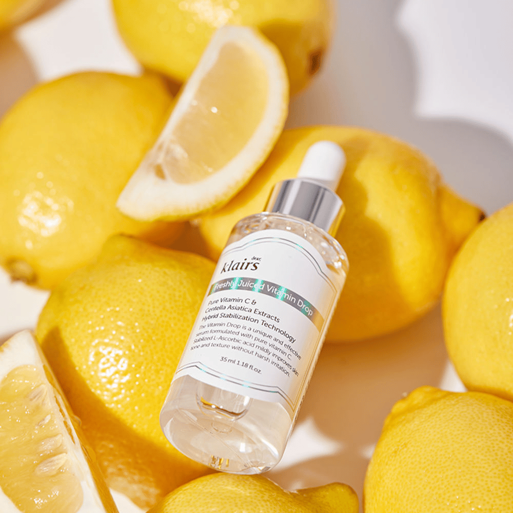 Klairs Freshly Juiced Vitamin Drop μπουκάλι ορού περιτριγυρισμένο από φρέσκα μισά λεμόνια και φέτες λεμονιού σε φωτεινή επιφάνεια, αναδεικνύει τα συστατικά.