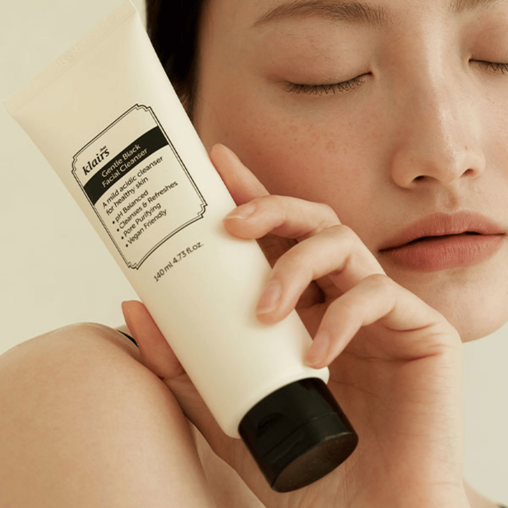 Ένα άτομο κρατά έναν σωλήνα Klairs Gentle Black Facial Cleanser στο μάγουλό του, με κλειστά μάτια και φυσικό τόνο δέρματος να φαίνεται στο φόντο.