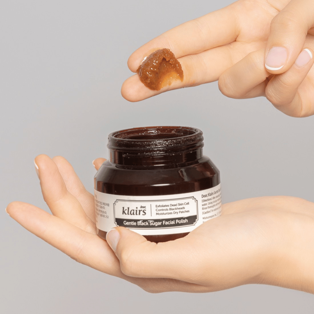 Χέρια κρατούν ένα ανοιχτό βάζο από Klairs Gentle Black Sugar Facial Polish και δείχνουν το προϊόν σε ένα δάχτυλο, σε γκρι φόντο.