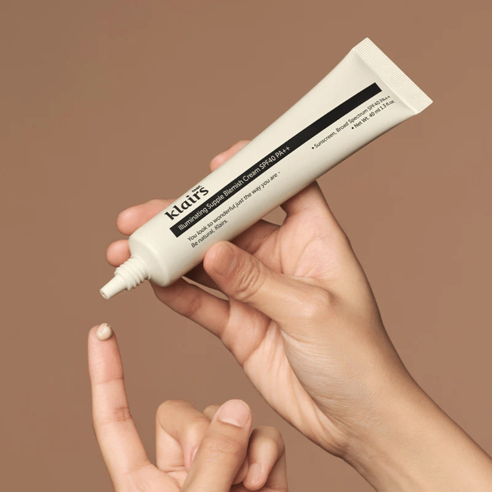 Ένα χέρι κρατάει ένα σωληνάριο Klairs Illuminating Supple Blemish Cream SPF40 PA++ και πιέζει μια μικρή ποσότητα κρέμας σε ένα δάχτυλο, σε καφέ φόντο.