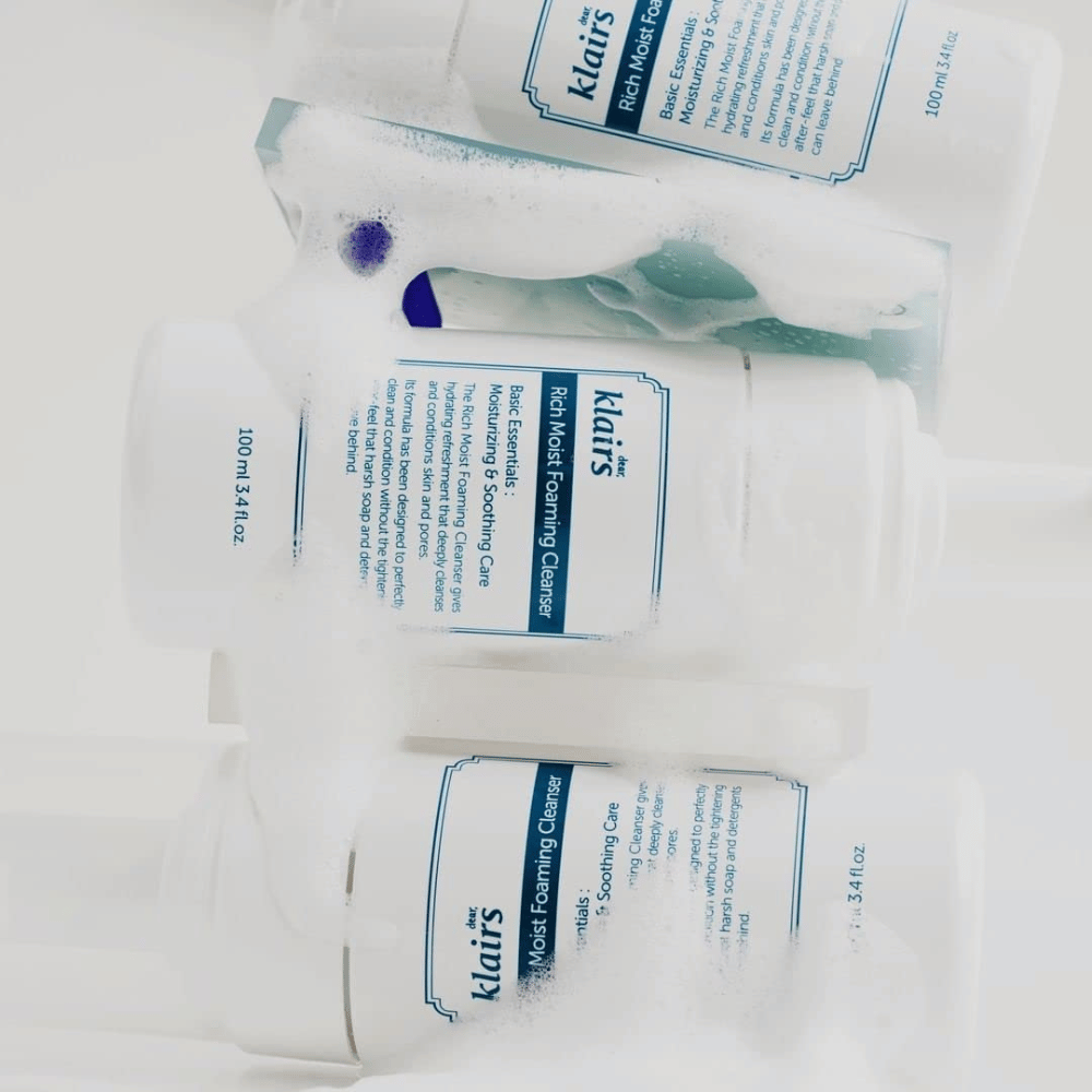 Τρία μπουκάλια Klairs Rich Moist Foaming Cleanser καλυμμένα με αφρό και σταγόνες νερού, τοποθετημένα σε λευκή επιφάνεια.