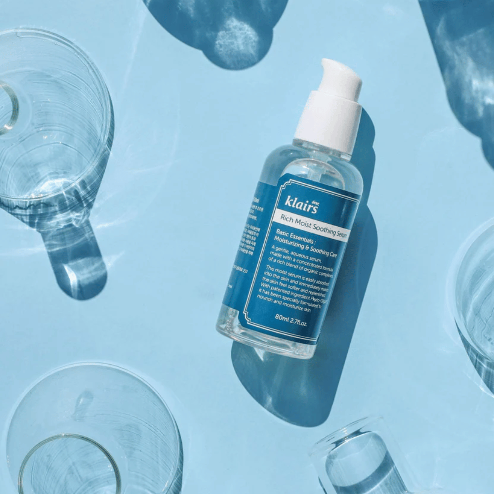 Ένα μπουκάλι Klairs Rich Moist Soothing Serum ανάμεσα σε ποτήρια νερού με αντανακλάσεις σε μπλε φόντο.