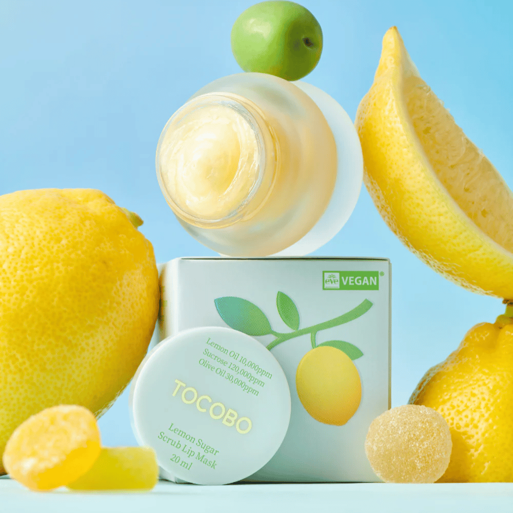 Βάζο με Tocobo Lemon Sugar Scrub Lip Mask τοποθετημένο ανάμεσα σε λεμόνια και συσκευασία, χαρακτηρισμένο ως φιλικό προς τους βίγκαν.
