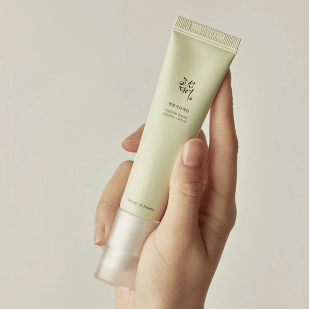 Ένα χέρι κρατάει το Beauty of Joseon Light On Serum με Centella και βιταμίνη C, σε ένα ανοιχτό πράσινο σωληνάριο με μινιμαλιστικό σχεδιασμό σε ουδέτερο φόντο.