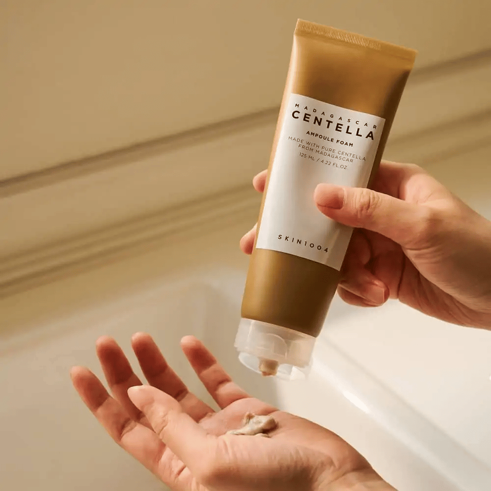Το προϊόν στην εικόνα είναι "Madagascar Centella Ampoule Foam" από SKIN1004. Είναι ένα καθαριστικό προσώπου σε μορφή αφρού, που παρασκευάζεται από Centella από τη Μαδαγασκάρη. Περιέχει 125 ml / 4.22 fl.oz. Ένα χέρι πιέζει μια μικρή ποσότητα προϊόντος για χρήση.