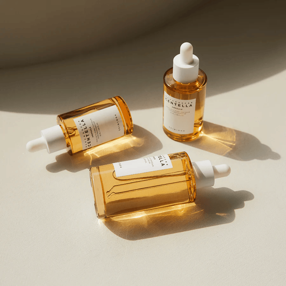 Η εικόνα δείχνει τρία μπουκάλια του "Madagascar Centella Asiatica 100 Ampoule". Είναι τοποθετημένα σε μια επιφάνεια όπου το ηλιακό φως δημιουργεί μια όμορφη αντανάκλαση μέσα από το χρυσό περιεχόμενο των μπουκαλιών. Το προϊόν φαίνεται να είναι μέρος μιας σειράς περιποίησης δέρματος που χρησιμοποιεί centella asiatica, ένα συστατικό γνωστό για τις καταπραϋντικές του ιδιότητες. Ο σχεδιασμός των μπουκαλιών είναι απλός και μοντέρνος, υποδηλώνοντας ότι πρόκειται για μια premium σειρά περιποίησης δέρματος.