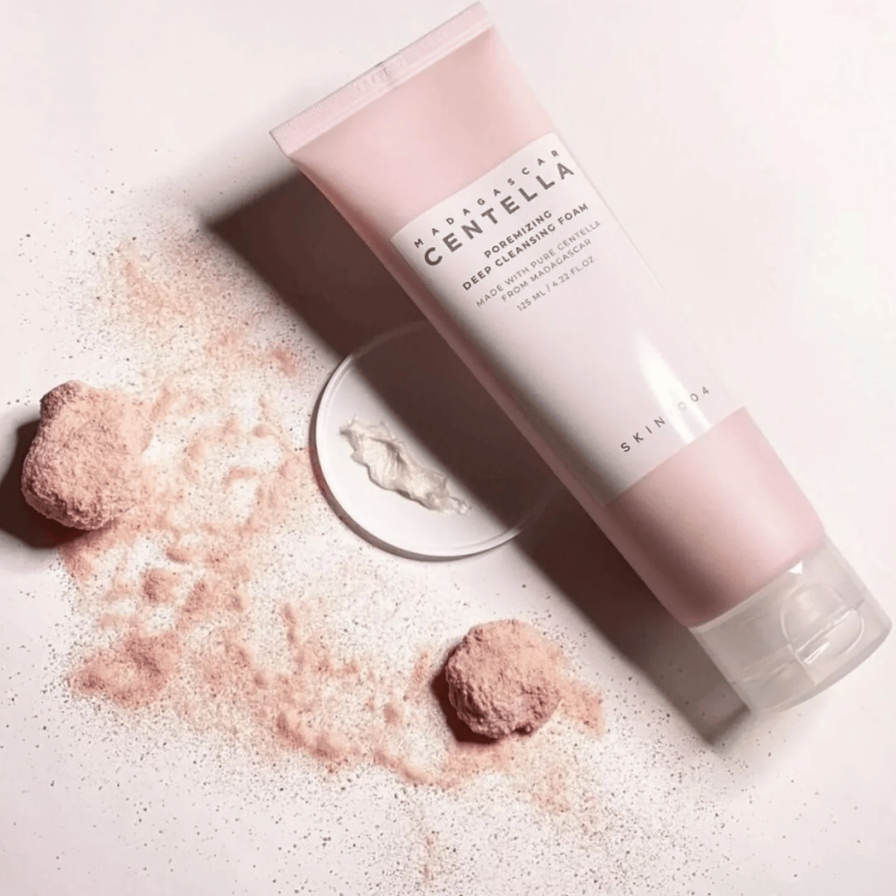 Madagascar Centella Poremizing Deep Cleansing Foam παρουσιάζεται σε ροζ σωληνάριο. Τα σωληνάρια βρίσκονται σε ανοιχτόχρωμη επιφάνεια μαζί με ροζ σκόνη που δίνει αίσθηση καθαριότητας και φρεσκάδας. Το προϊόν είναι σχεδιασμένο για βαθύ καθαρισμό του δέρματος με τη βοήθεια καθαρής centella από τη Μαδαγασκάρη.