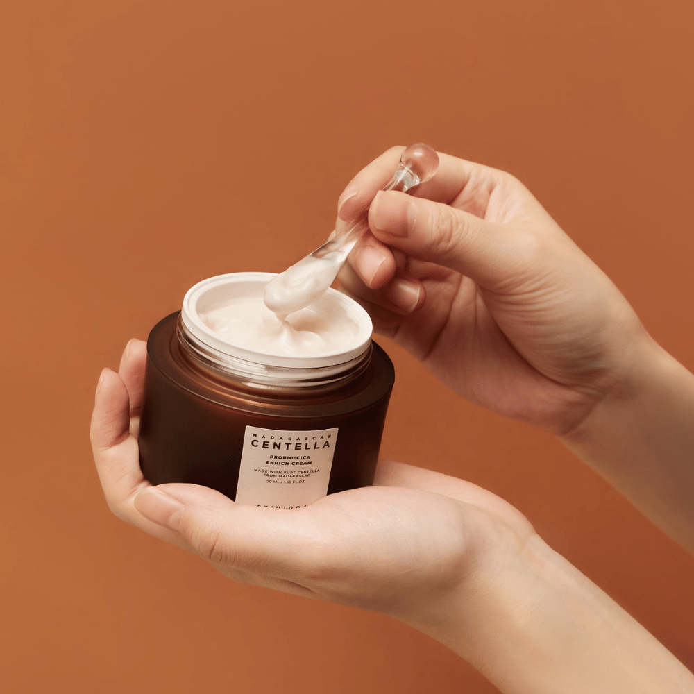 Χέρια που κρατούν ένα ανοιχτό βάζο από Madagascar Centella Probio-Cica Enrich Cream και μια σπάτουλα με κρέμα σε ένα ομοιόμορφο φόντο σε χρώμα τερακότας.
