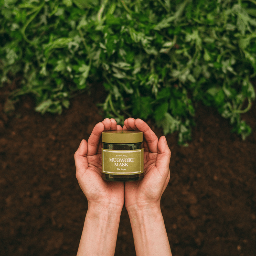 Χέρια που κρατούν το I'm From Mugwort Mask σε φόντο χώματος και φρέσκων πράσινων βοτάνων, τονίζοντας τα φυσικά συστατικά του προϊόντος.