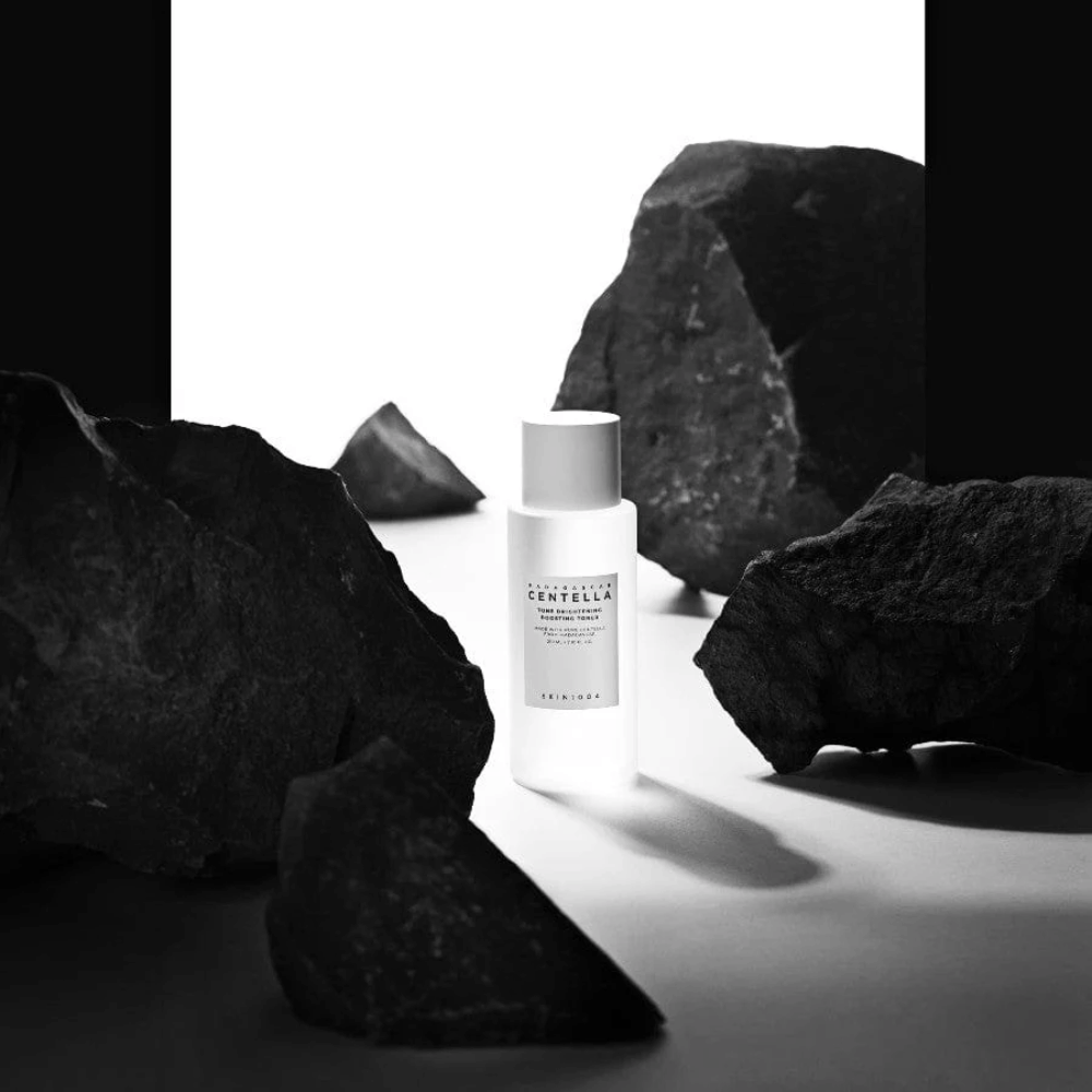 Λευκό μπουκάλι SKIN1004 Madagascar Centella Tone Brightening Boosting Toner  τοποθετημένο δραματικά ανάμεσα σε μεγάλες μαύρες πέτρες με λευκό φόντο.