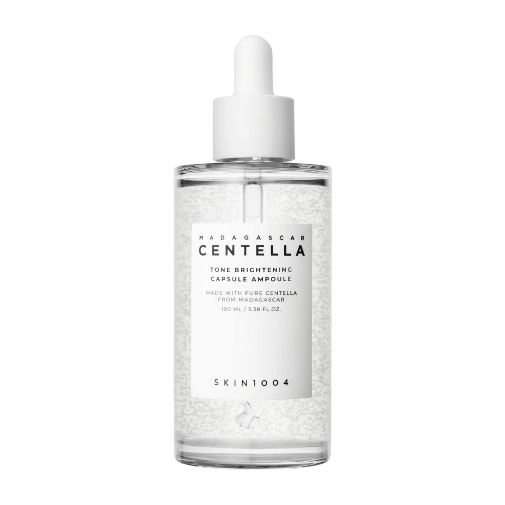 Η εικόνα δείχνει ένα μπουκάλι του "SKIN1004 Madagascar Centella Tone Brightening Capsule Ampoule". Είναι ένα διαφανές μπουκάλι με λευκό εφαρμογέα σταγόνων. Στην ετικέτα αναφέρεται ότι το προϊόν περιέχει καθαρά συστατικά centella από τη Μαδαγασκάρη. Το περιεχόμενο της φιάλης φαίνεται να έχει μικρές κάψουλες ή σωματίδια, κάτι που μπορεί να είναι μέρος των φωτεινών ιδιοτήτων περιποίησης της επιδερμίδας του. Η φιάλη περιέχει 100 ml / 3.38 fl.oz. του προϊόντος.