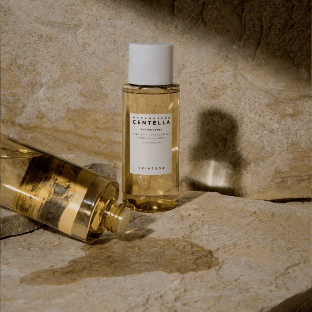 Η εικόνα δείχνει ένα μπουκάλι "Madagascar Centella Toning Toner" από SKIN1004. Είναι ένα διαφανές μπουκάλι γεμάτο με ένα κιτρινωπό υγρό, πιθανότατα έναν τόνερ για περιποίηση δέρματος. Αναφέρεται ότι είναι φτιαγμένο με καθαρή centella από τη Μαδαγασκάρη και το περιεχόμενο είναι 210 ml / 7.10 fl.oz. Το μπουκάλι και ένα πλάγιο μπουκάλι είναι τοποθετημένα σε μια πετρώδη επιφάνεια, που δίνει μια φυσική και μινιμαλιστική αίσθηση.