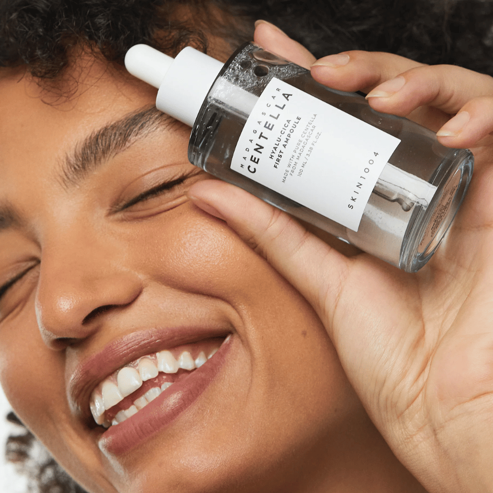 Χαμογελαστή γυναίκα κρατάει Skin1004 Madagascar Centella Hyalu-Cica First Ampoule δίπλα στο πρόσωπό της.