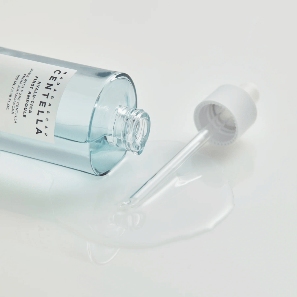 Κοντινή λήψη ανοιχτού μπουκαλιού Skin1004 Madagascar Centella Hyalu-Cica First Ampoule με πιπέτα ξαπλωμένη και ορό απλωμένο σε λευκή επιφάνεια.