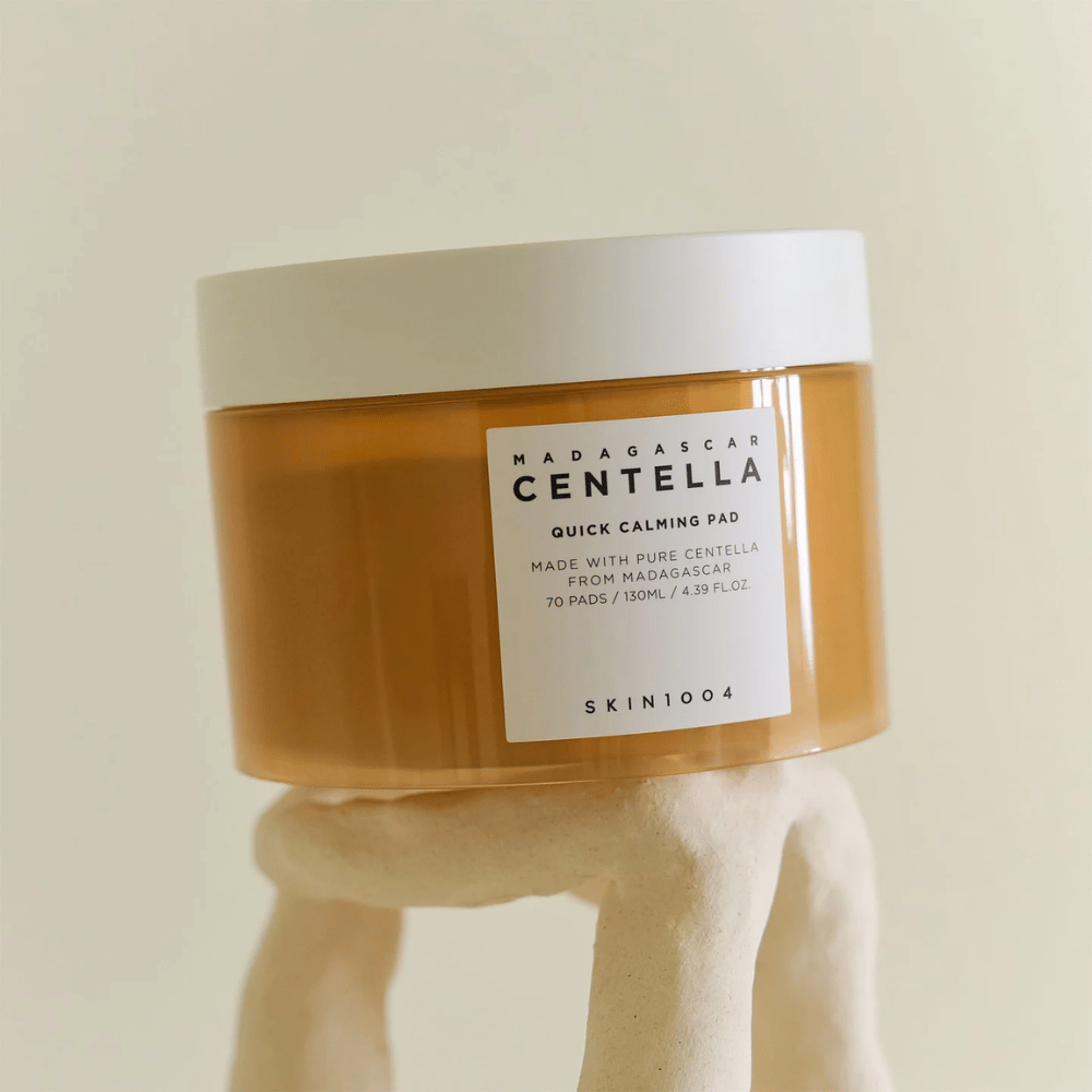 Δοχείο με SKIN1004 Madagascar Centella Quick Calming Pad τοποθετημένο σε γλυπτική λευκή βάση – κεχριμπαρένιο δοχείο με λευκή ετικέτα και καπάκι.