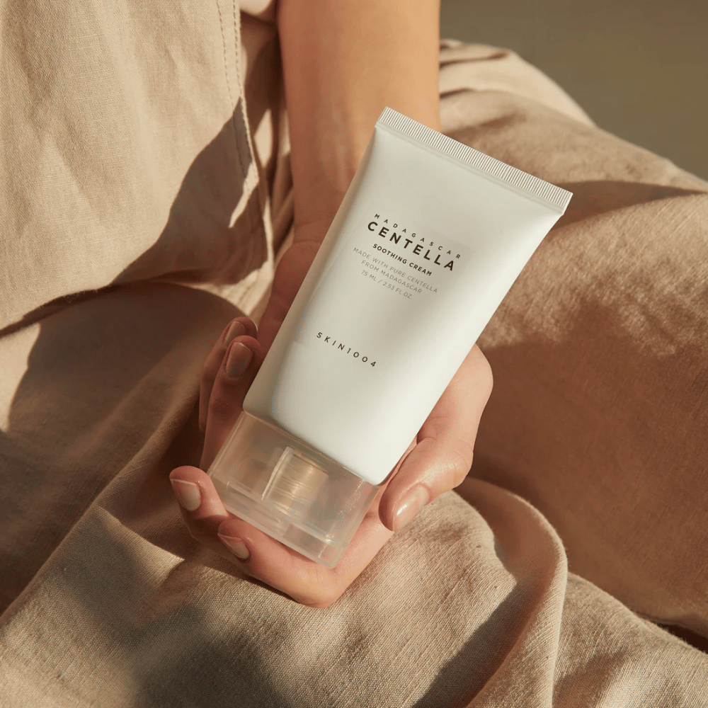 Ένα άτομο κρατάει έναν λευκό σωλήνα από Madagascar Centella Soothing Cream από SKIN1004. Ο σωλήνας έχει διάφανο καπάκι και κείμενο που περιγράφει το προϊόν ως καταπραϋντικό και κατασκευασμένο με καθαρή Centella. Το άτομο φοράει ανοιχτόχρωμα ρούχα.