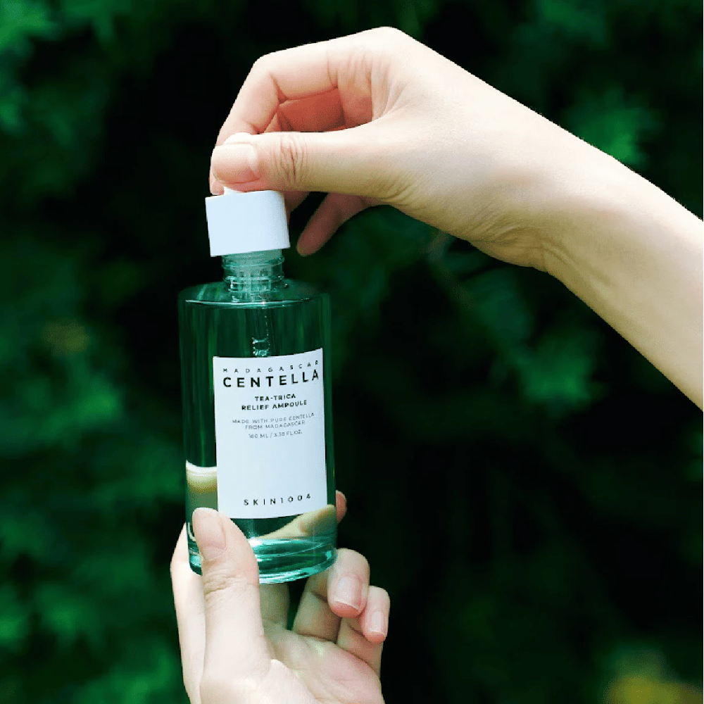 Άτομο ανοίγει SKIN1004 Madagascar Centella Tea-Trica Relief Ampoule – πράσινο γυάλινο μπουκάλι με λευκή ετικέτα κρατιέται στα χέρια μπροστά από πράσινο φόντο.
