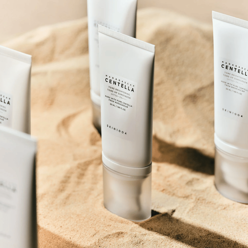 Πολλοί σωλήνες με Madagascar Centella Tone Brightening Tone-Up Sunscreen στέκονται στην άμμο. Ο σωλήνας είναι λευκός με μαύρο κείμενο και περιέχει 50 ml. Το προϊόν έχει SPF 50+ PA++++ και είναι κατασκευασμένο με καθαρή Centella από τη Μαδαγασκάρη.