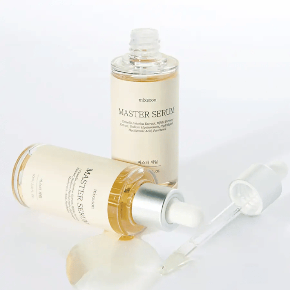 Δύο μπουκάλια Mixsoon Master Serum, το ένα όρθιο και το άλλο ξαπλωμένο με ανοιχτή πιπέτα, όπου ο ορός έχει χυθεί σε μια λευκή επιφάνεια.
