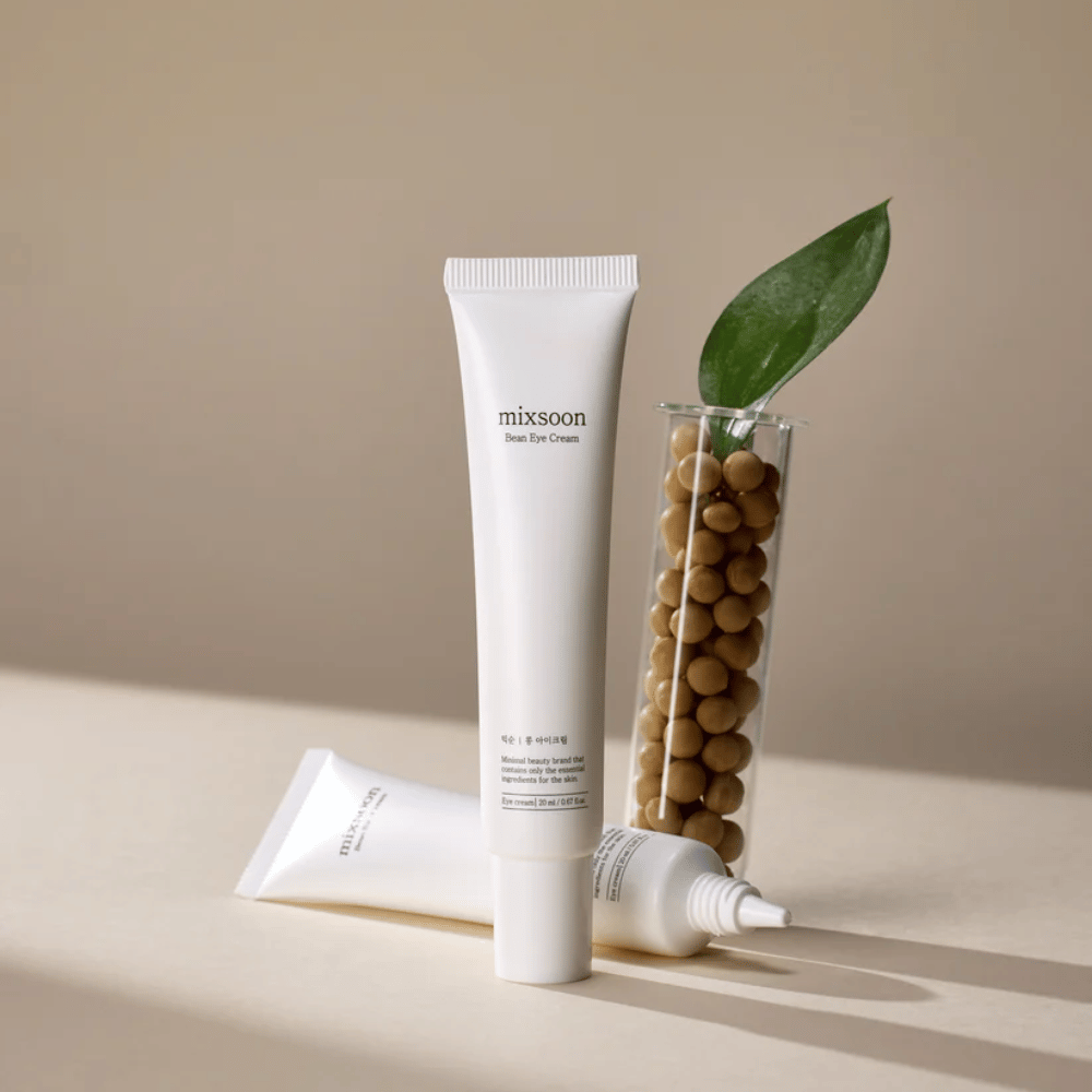 Δύο σωλήνες Mixsoon Bean Eye Cream τοποθετημένες δίπλα σε έναν δοκιμαστικό σωλήνα γεμάτο με σόγια και ένα πράσινο φύλλο φυτού, σε μια μπεζ επιφάνεια με παιχνίδια σκιών.