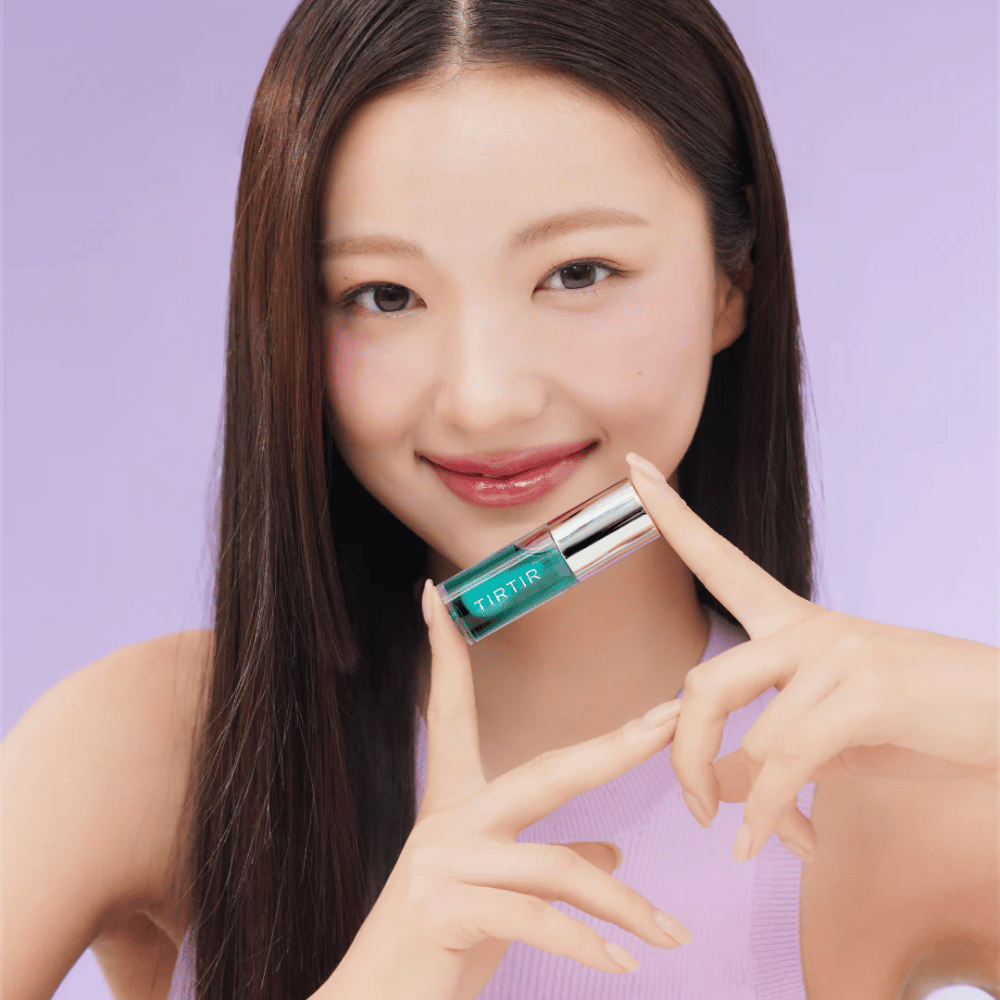 Γυναίκα με μακριά, ίσια μαλλιά κρατάει TIRTIR My Glow Lip Oil και δείχνει τα γυαλιστερά, ροζ χείλη της σε μοβ φόντο.