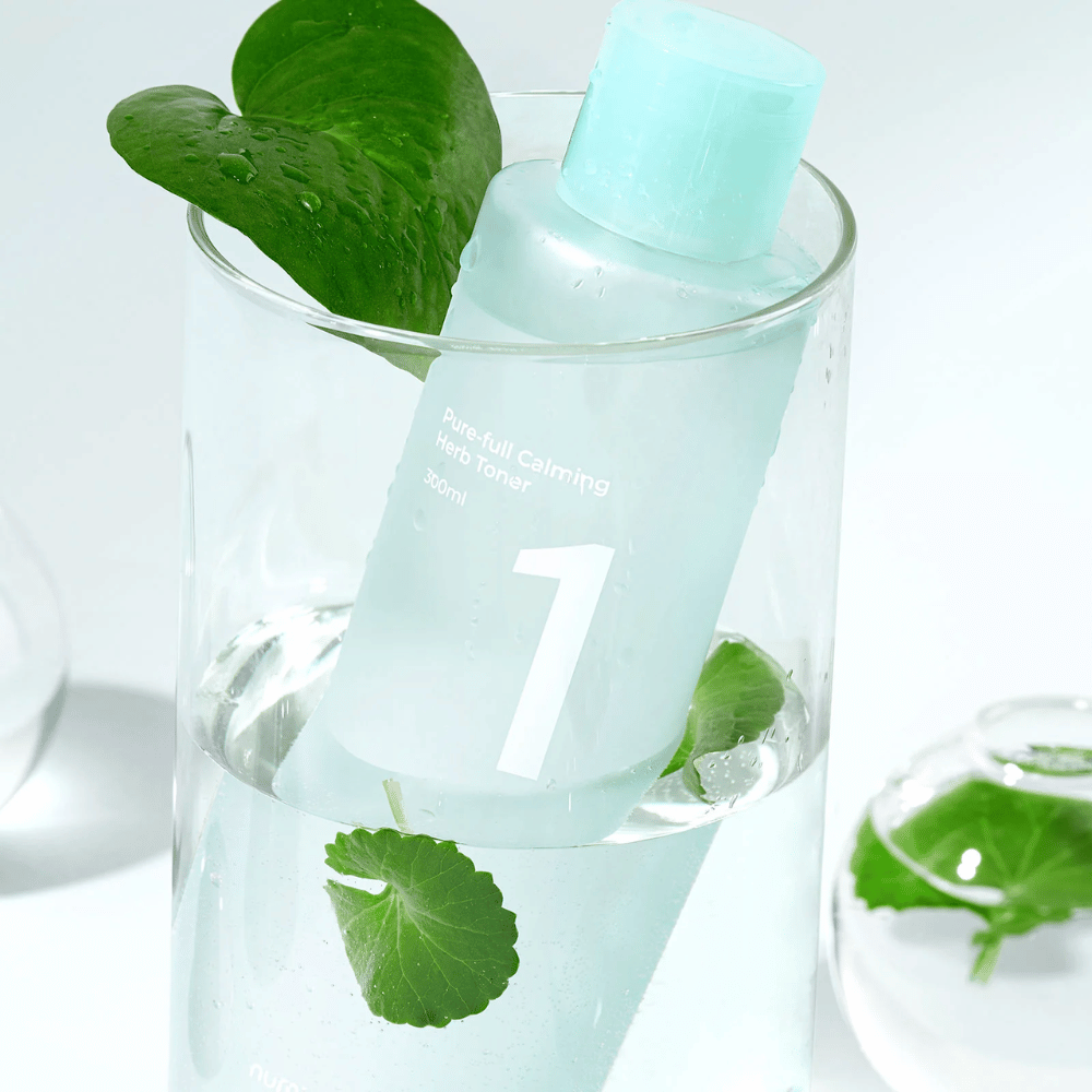 Ένα μπουκάλι με No.1 Pure-Full Calming Herb Toner τοποθετημένο σε ένα ποτήρι με νερό και πράσινα φύλλα. Το μπουκάλι έχει ένα ανοιχτό μπλε καπάκι και μια ετικέτα με το κείμενο "Pure-full Calming Herb Toner" καθώς και "300ml". Το προϊόν φαίνεται φρέσκο με νερό και βλάστηση στο παρασκήνιο, το οποίο δίνει μια καταπραϋντική και φυσική αίσθηση.