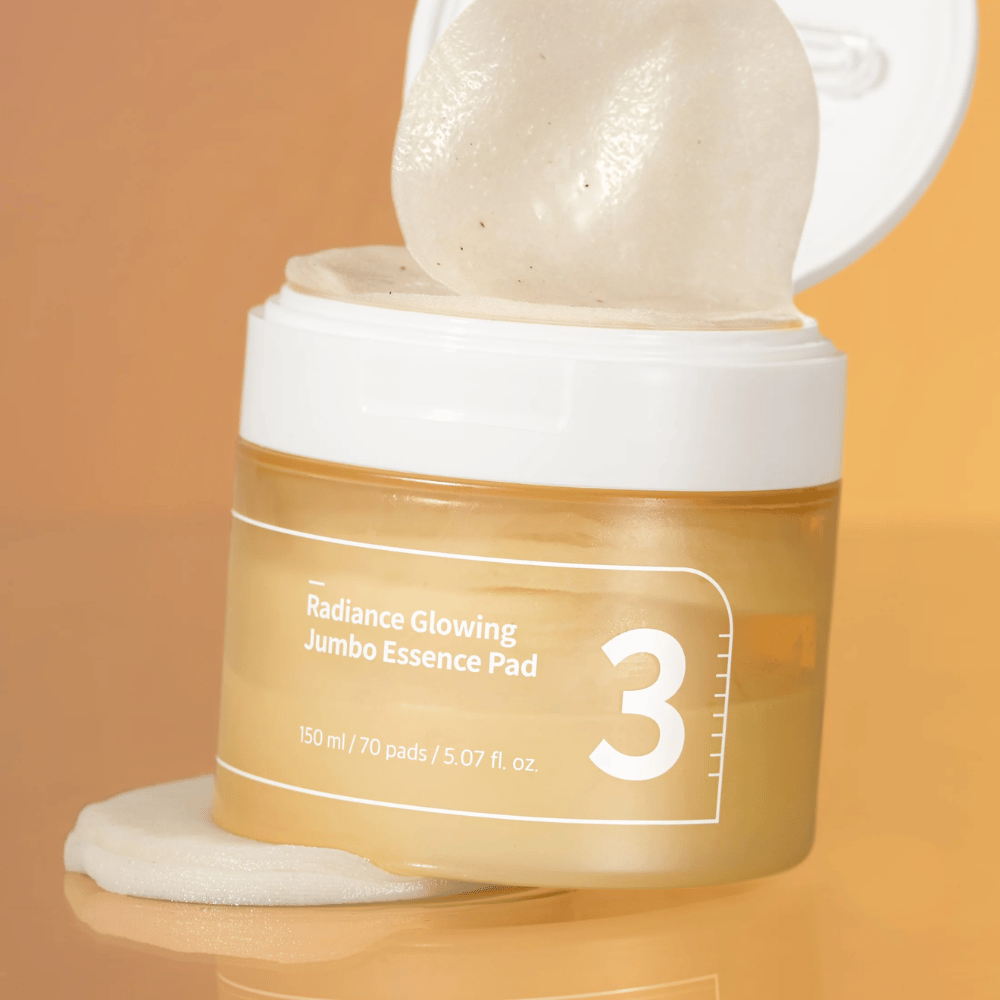 Ανοιχτό δοχείο με numbuzin No. 3 Radiance Glowing Jumbo Essence Pad, ένα pad φαίνεται γερμένο στο χείλος, φόντο σε πορτοκαλί απόχρωση.