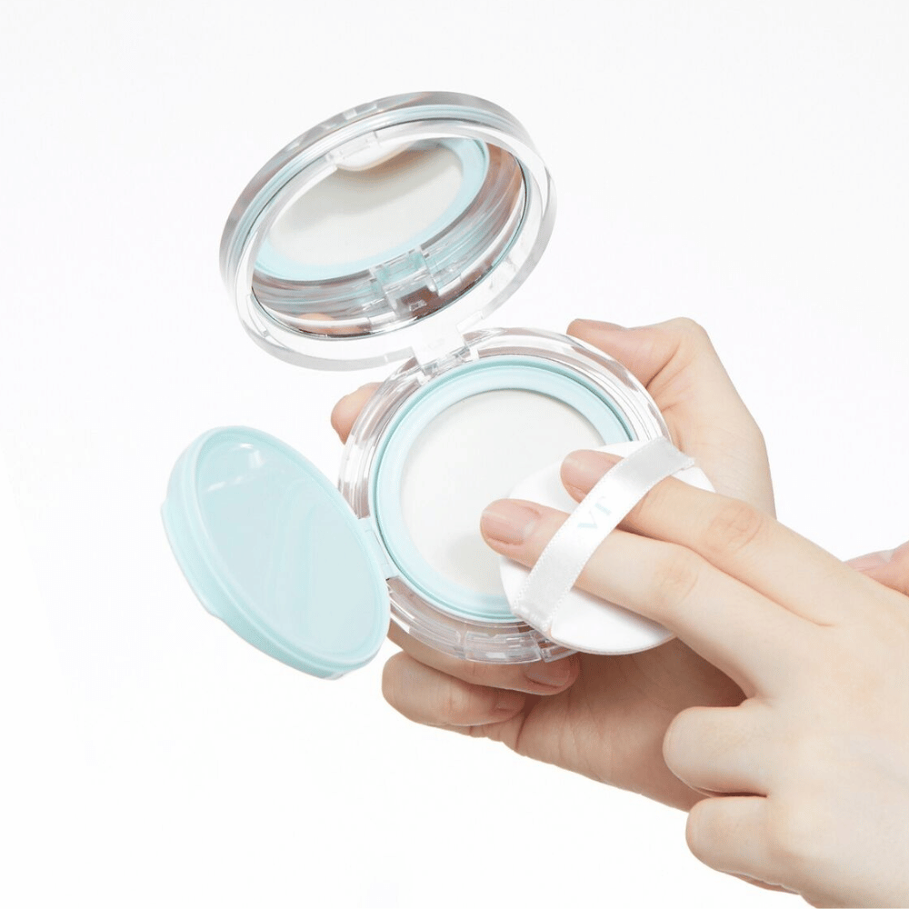 Ένα χέρι κρατά ένα ανοιχτό βάζο με VT PDRN Essence Glow Sun Pact. Το πούφ είναι έτοιμο να εφαρμόσει την πούδρα, η οποία έχει λευκή επιφάνεια και μπλε περίγραμμα γύρω από το βάζο.