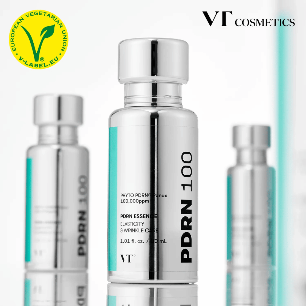 Αρκετά ασημί μπουκάλια της VT PDRN 100 Essence είναι παρατεταγμένα, με σαφή σήμανση για 100,000 ppm PDRN και εστίαση στην ελαστικότητα και φροντίδα ρυτίδων. Στην κορυφή φαίνεται το λογότυπο της VT Cosmetics και ένα V-label για χορτοφαγικό περιεχόμενο.